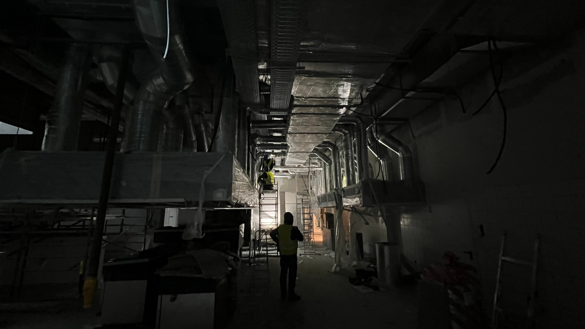 Montaż instalacji wentylacyjnej w budynku komercyjnym. Widoczne kanały wentylacyjne pod sufitem i pracownicy na drabinach w żółtych kamizelkach. Słabe oświetlenie.