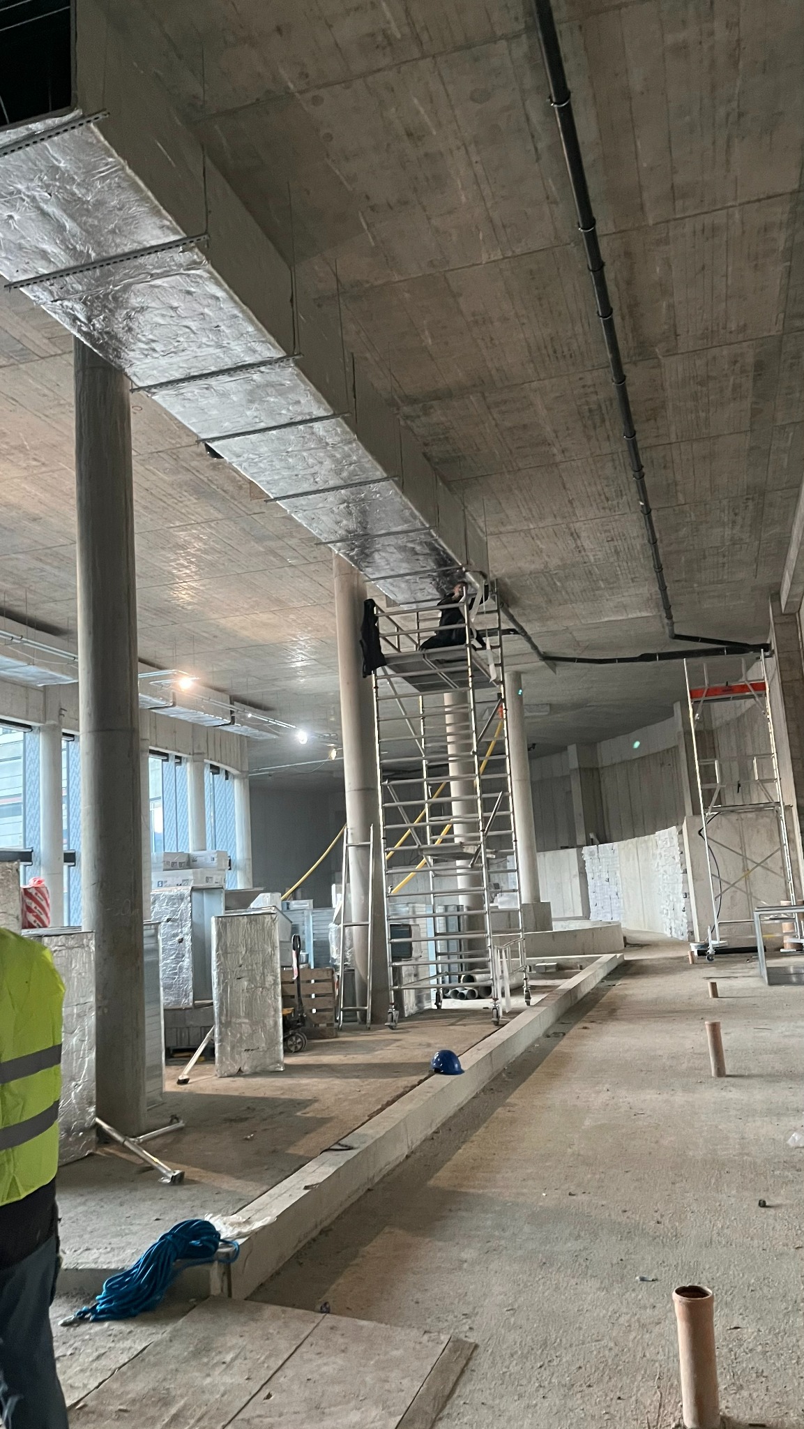 Montaż instalacji wentylacyjnej w surowym wnętrzu budynku komercyjnego. Widoczne kanały wentylacyjne pod sufitem, rusztowania i pracownik w trakcie pracy.