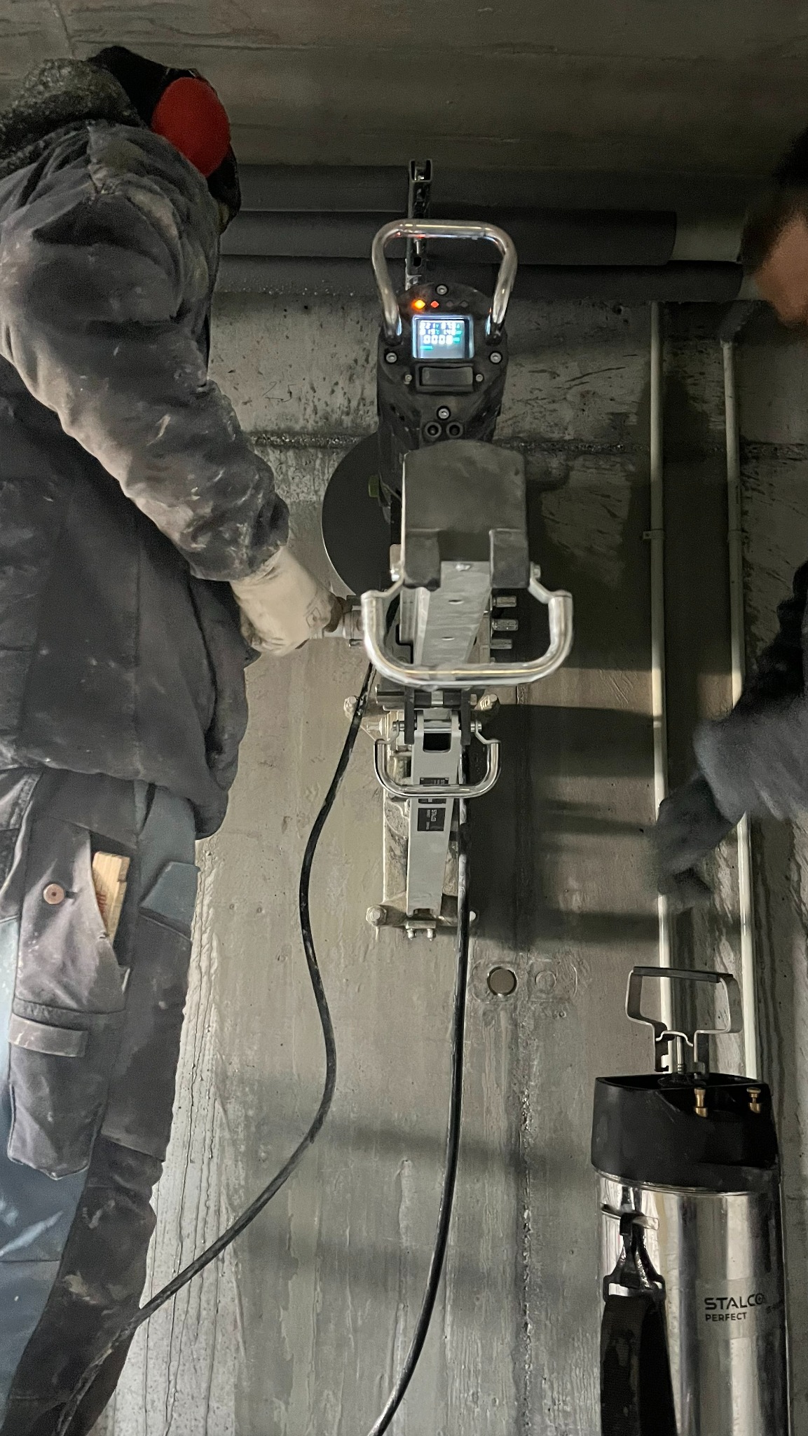 Pracownik w trakcie wykonywania precyzyjnego odwiertu w betonowej ścianie za pomocą specjalistycznego wiertła diamentowego z odsysaniem, widoczny zbiornik na wodę.