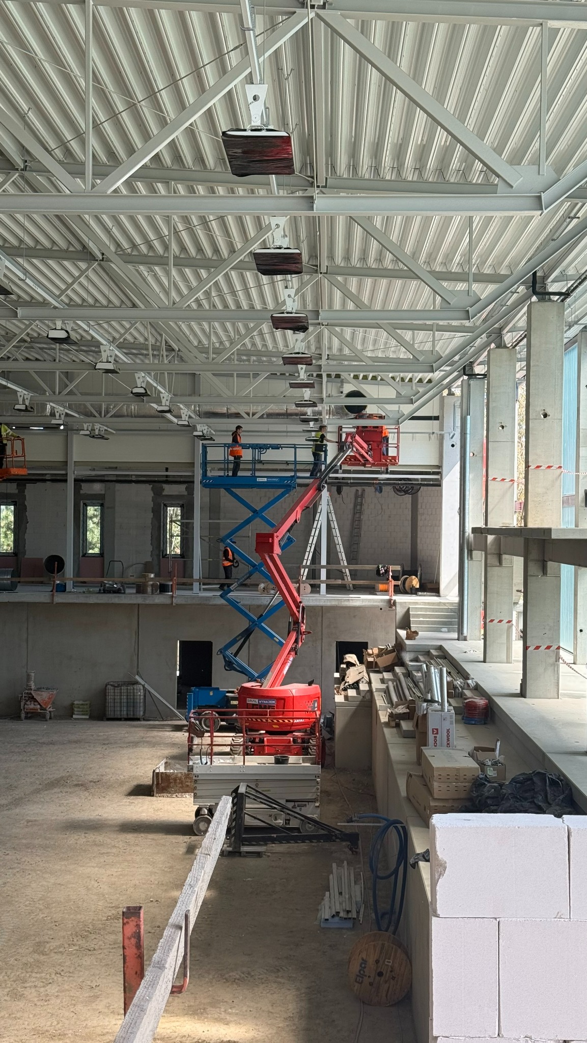 Montaż instalacji w hali: podnośnik z pracownikami przy suficie z widocznymi elementami wentylacji. Surowy stan, betonowe ściany, okna. Widok z dołu na konstrukcję.