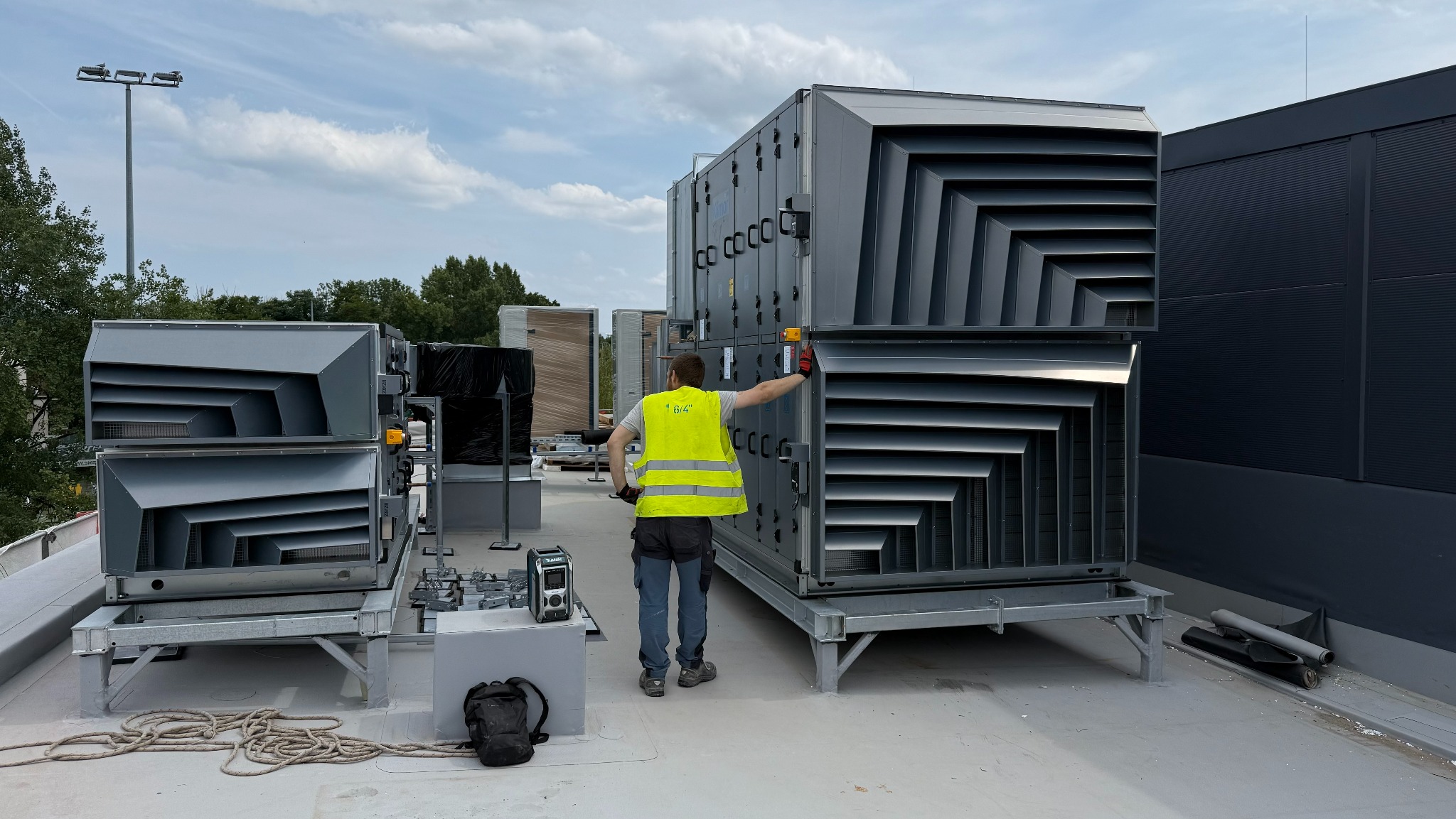 Instalacja rekuperacji na dachu budynku komercyjnego. Pracownik w kamizelce mierzy poziom. Widoczne elementy systemu wentylacyjnego i narzędzia.