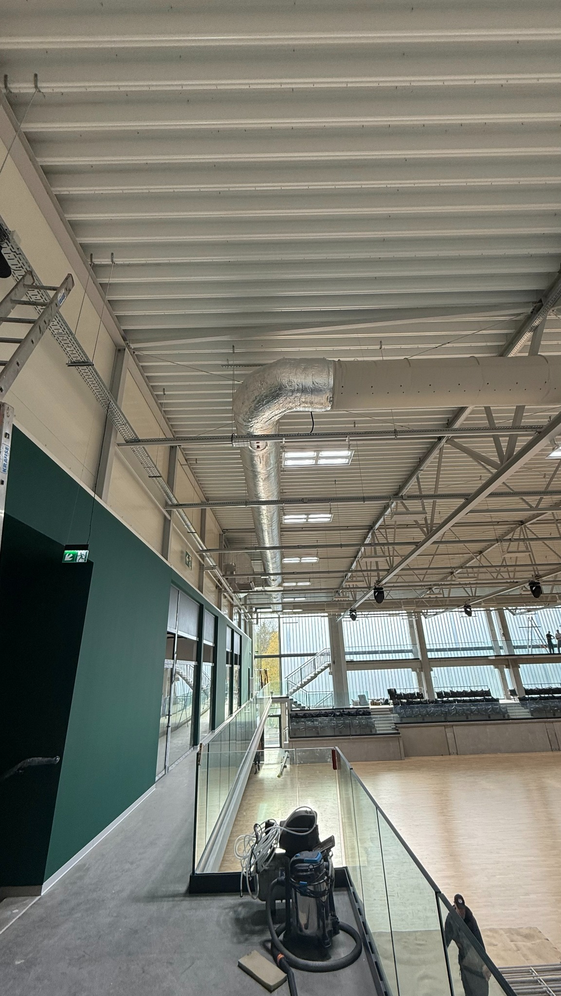 Wnętrze hali sportowej z widoczną instalacją wentylacyjną pod sufitem. Na pierwszym planie sprzęt czyszczący i fragment balustrady. Widoczne okna i konstrukcja dachu.