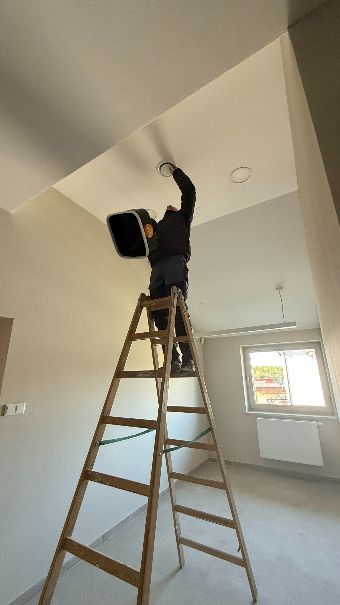 Instalator na drabinie montuje okrągły nawiewnik sufitowy, trzymając urządzenie pomiarowe Testo. Widoczne okno i grzejnik w jasnym pomieszczeniu.