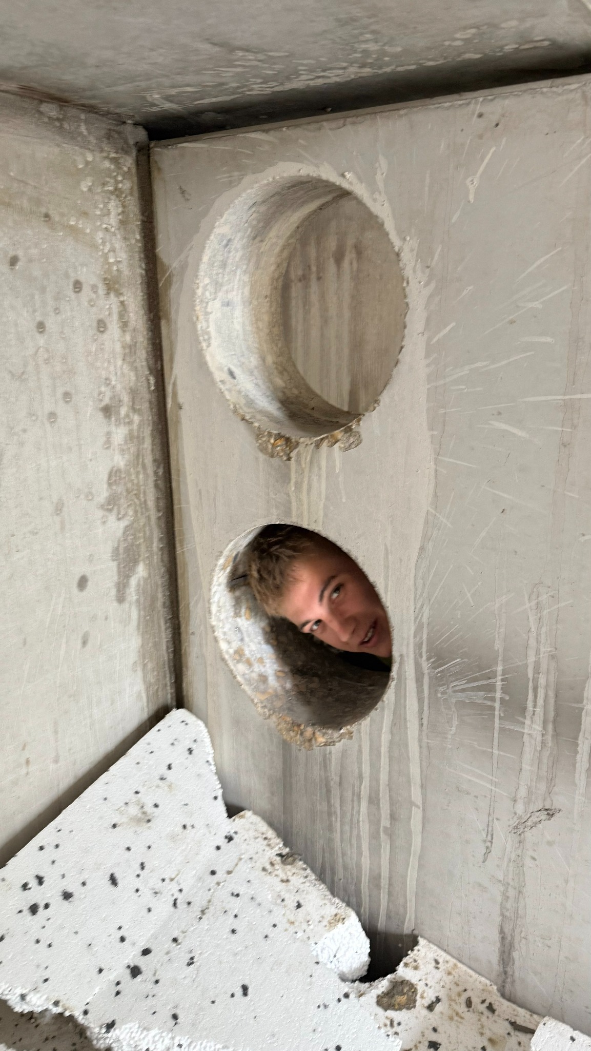 Betonowa ściana z dwoma okrągłymi otworami, z których jednego wygląda uśmiechnięta osoba. Widoczne ślady po wierceniu i fragmenty gruzu.