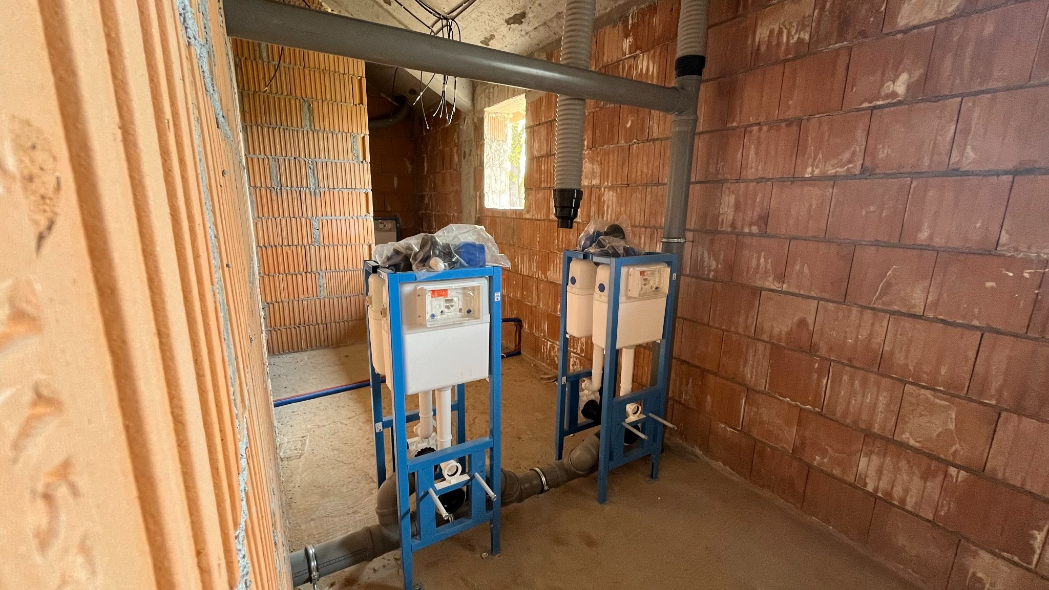 Dwa stelaże podtynkowe WC w surowym wnętrzu z ceglanymi ścianami. Widoczne rury kanalizacyjne i wentylacyjne. Instalacja w trakcie montażu, przed wykończeniem.