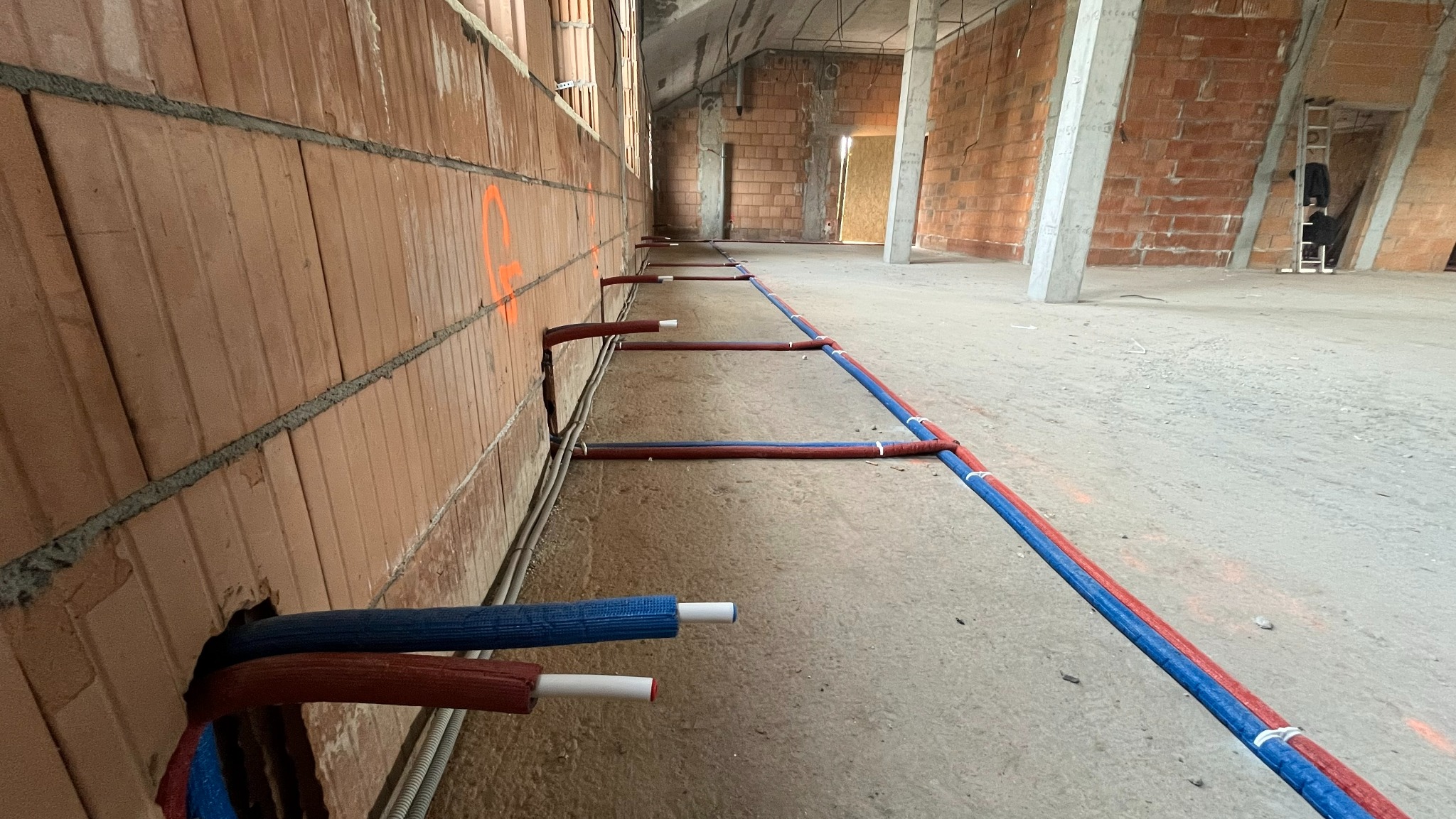 Instalacja rur grzewczych w surowym budynku z cegły. Rury w izolacji czerwonej i niebieskiej, prowadzone wzdłuż ściany, widoczne otwory w ścianie. W tle surowy stan wykończenia.