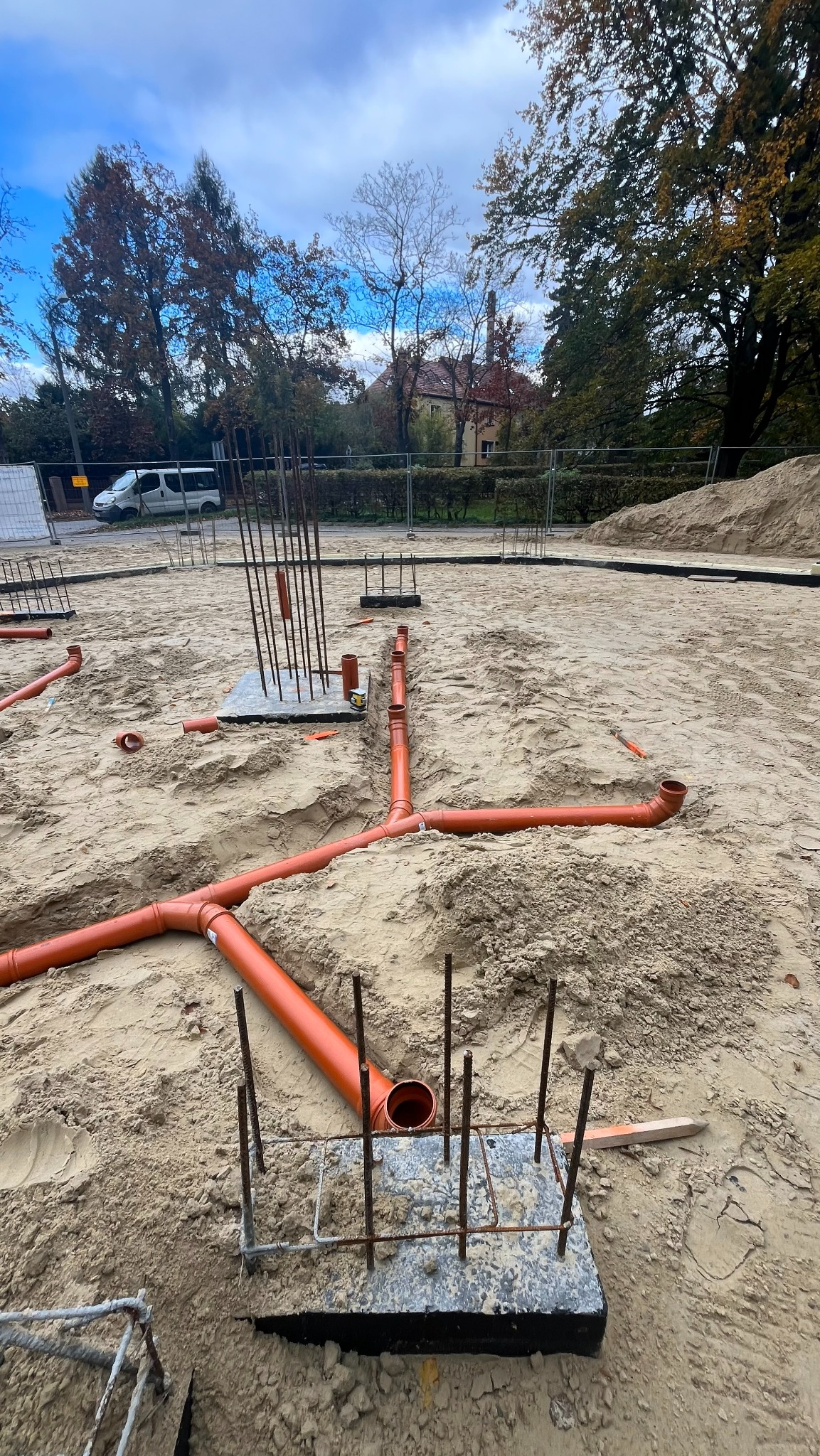 Pomarańczowe rury kanalizacyjne ułożone w piasku na placu budowy, widoczne zbrojenia fundamentów. Instalacja podposadzkowa w trakcie realizacji.