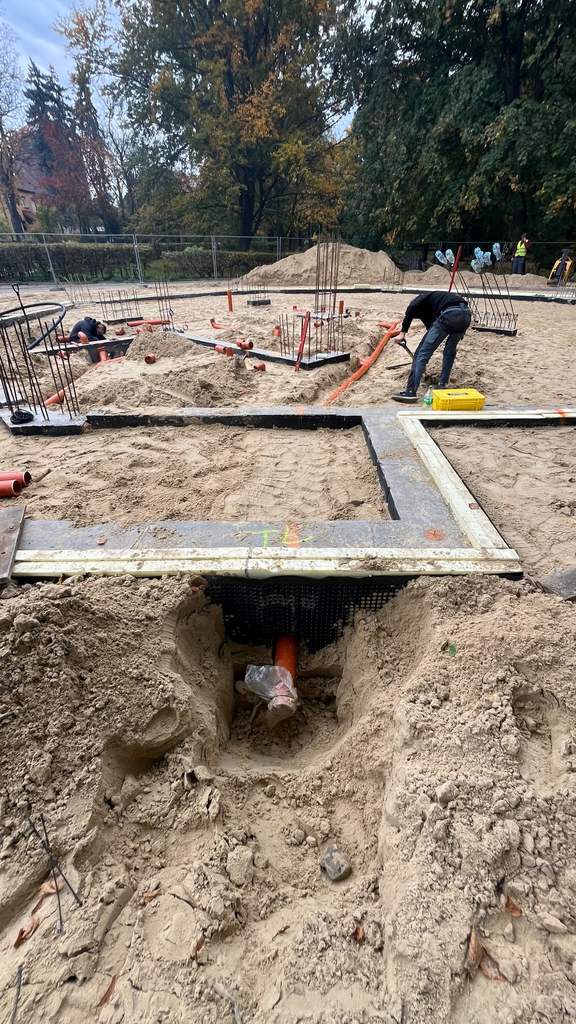 Instalacja rur w piasku na budowie, widoczne fundamenty i pracownicy. Pomarańczowe rury częściowo zakopane, żółta skrzynka narzędziowa w tle. Prace ziemne w toku.