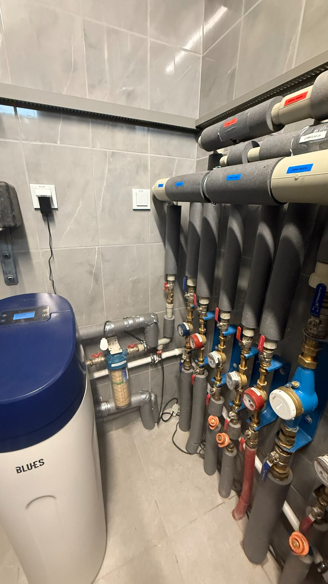 Węzeł cieplny z filtrem wody i rurami w izolacji, z licznymi zaworami i manometrami, na tle szarej, kafelkowanej ściany i podłogi. Widoczne oznaczenia na rurach.
