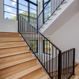 Dmytro Marienko - Nowoczesne schody drewniane z czarną, metalową balustradą w jasnym wnętrzu z dużymi oknami. Widok z dołu, podkreślający geometrię i minimalistyczny design.