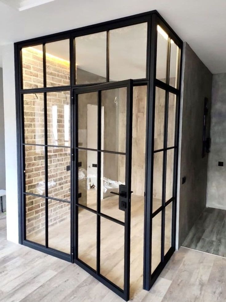 Loftowa ścianka działowa z czarnymi szprosami i przeszklonymi panelami, oddzielająca przestrzeń z ceglaną ścianą od szarego wnętrza. Otwarta, uchylona brama.