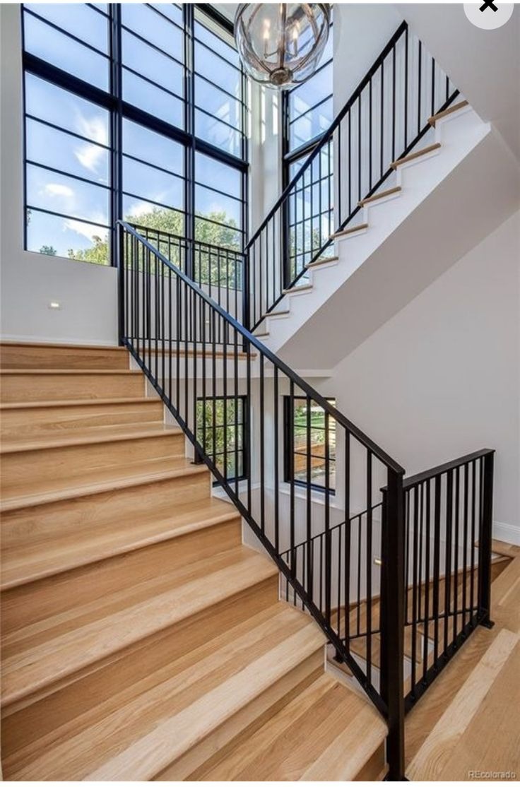 Nowoczesne schody drewniane z czarną, metalową balustradą w jasnym wnętrzu z dużymi oknami. Widok z dołu, podkreślający geometrię i minimalistyczny design.