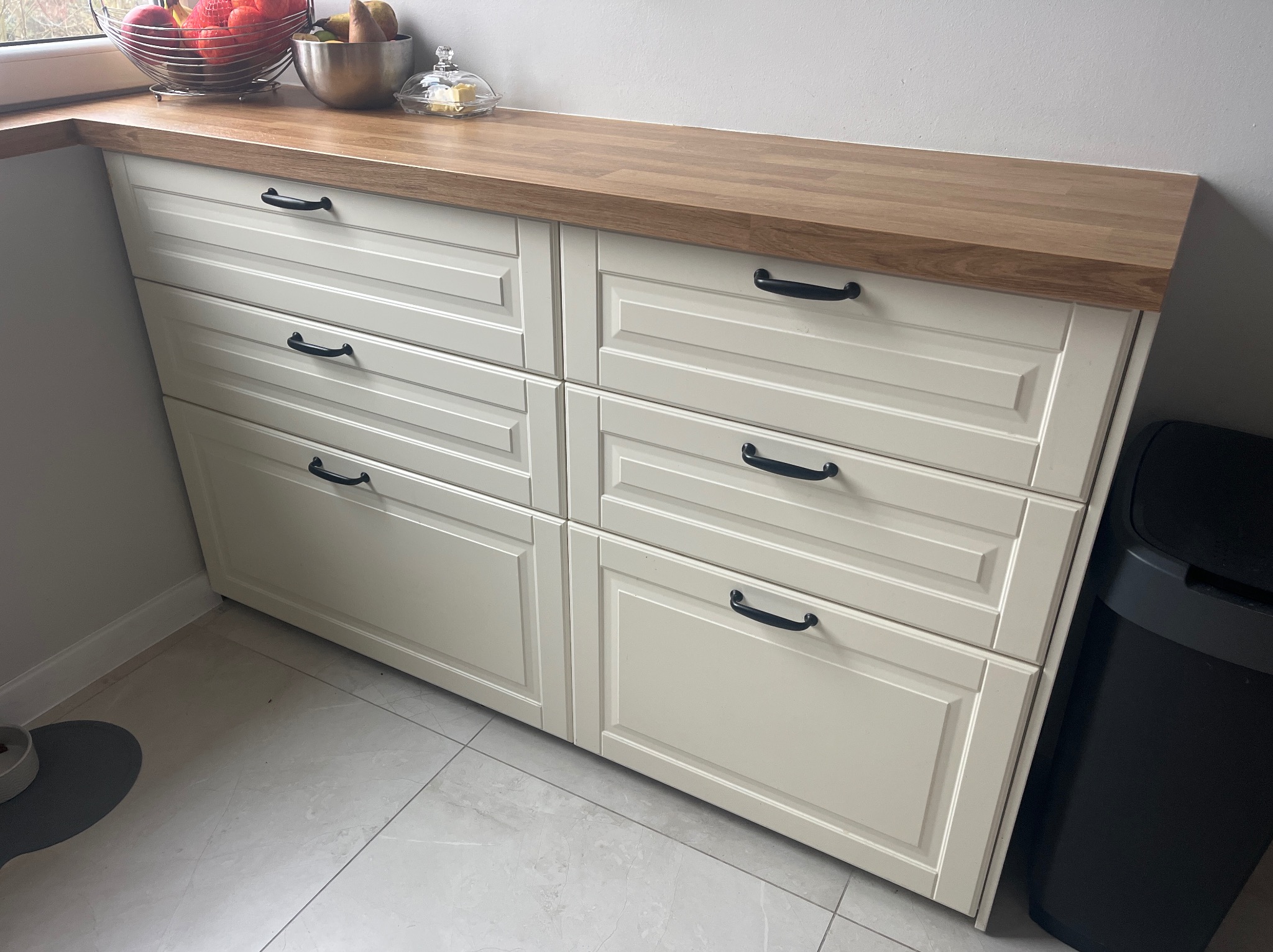 Beżowa komoda z drewnianym blatem i czarnymi uchwytami, ustawiona w rogu pomieszczenia. Na blacie owoce w metalowej misie i szklane naczynie z masłem.