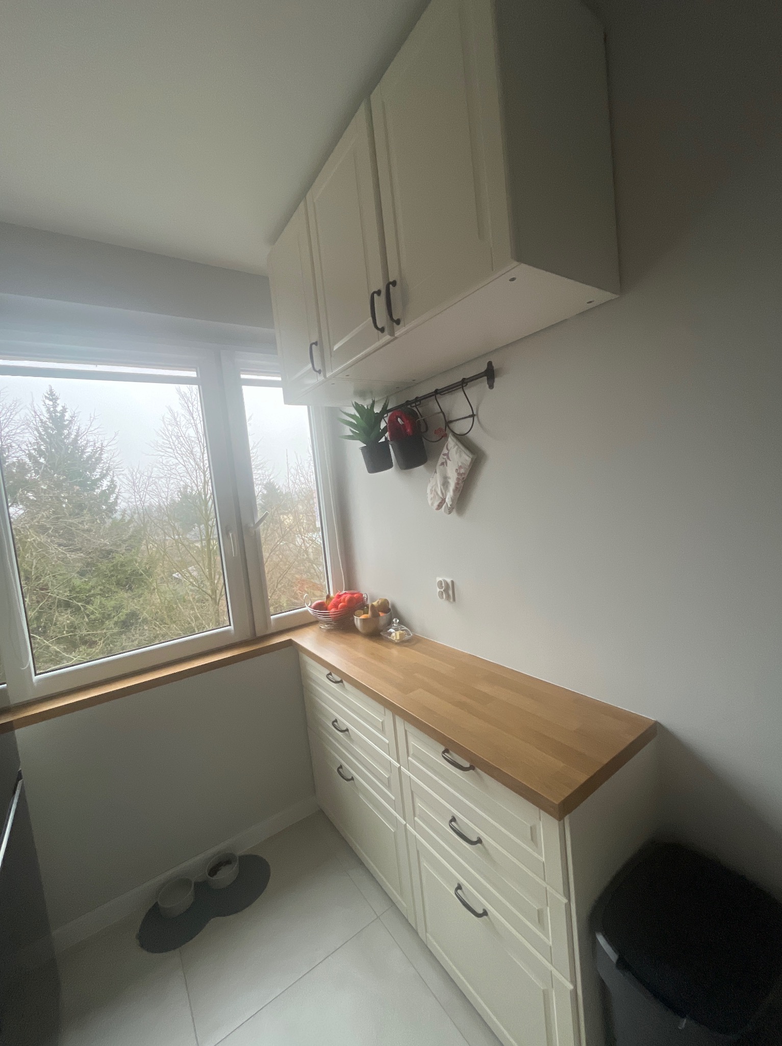 Jasna zabudowa kuchenna z drewnianym blatem w Łodzi, widok na okno z roślinami. Szafki wiszące z czarnymi uchwytami, reling z akcesoriami kuchennymi.