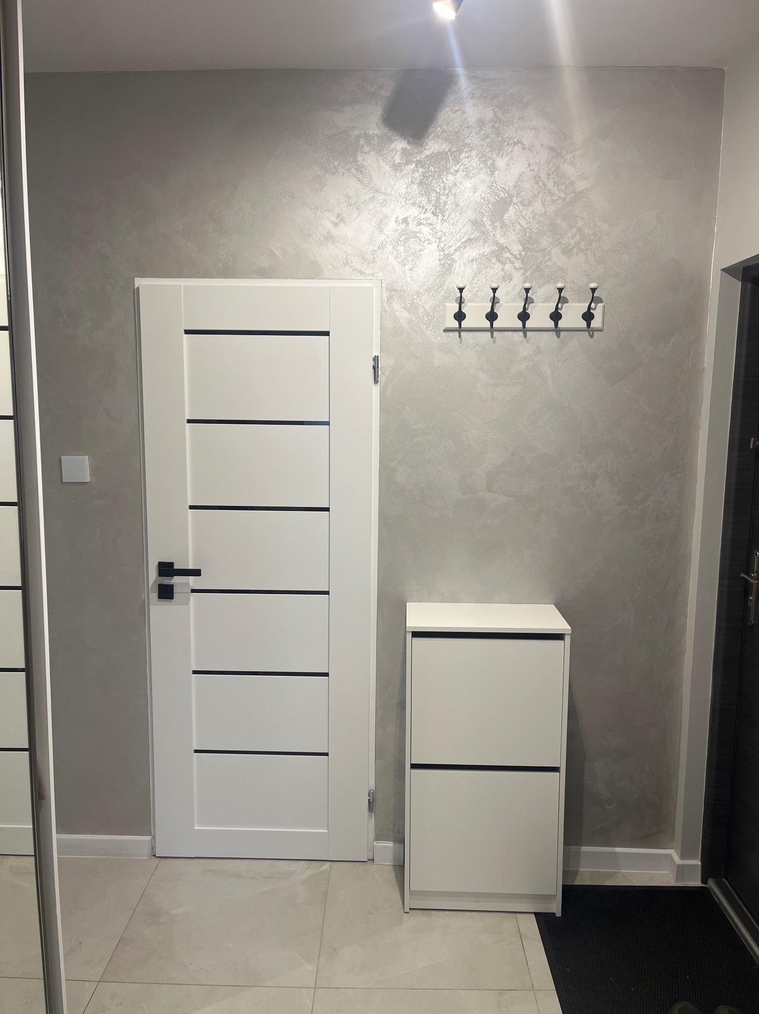 Nowoczesny przedpokój z białą szafką na buty, drzwiami z czarnymi akcentami i dekoracyjną szarą ścianą z wieszakiem. Minimalistyczny design.