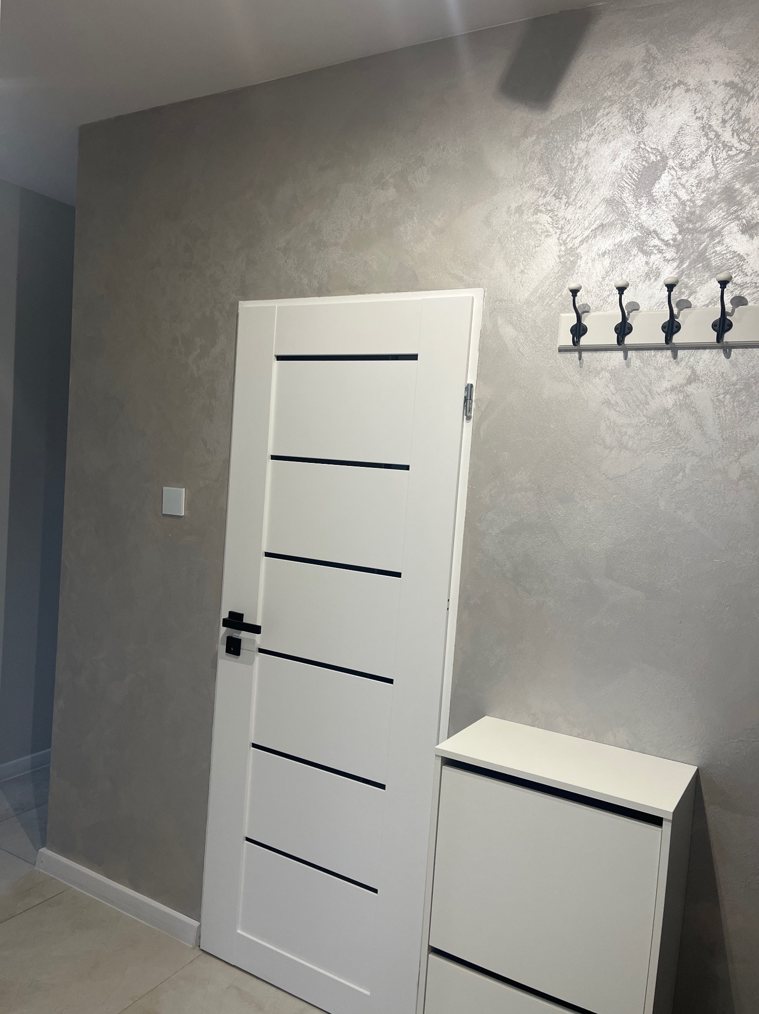 Białe drzwi z czarnymi akcentami, szafka na buty i wieszak na tle ściany z efektem dekoracyjnym w odcieniach szarości. Nowoczesne wykończenie wnętrza.