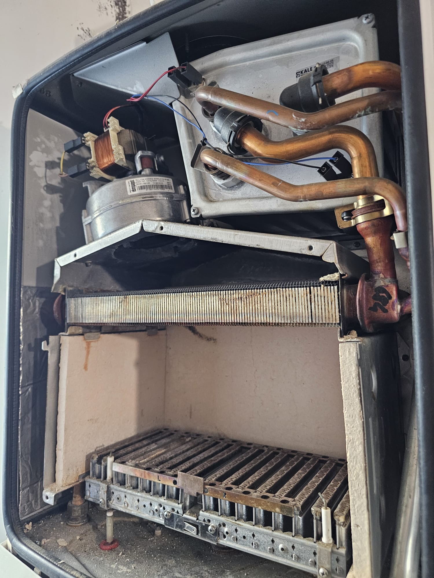 Otwarty piec gazowy z widocznymi elementami wewnętrznymi: rurami miedzianymi, palnikiem i elektroniką. Widoczne ślady użytkowania i zabrudzenia.