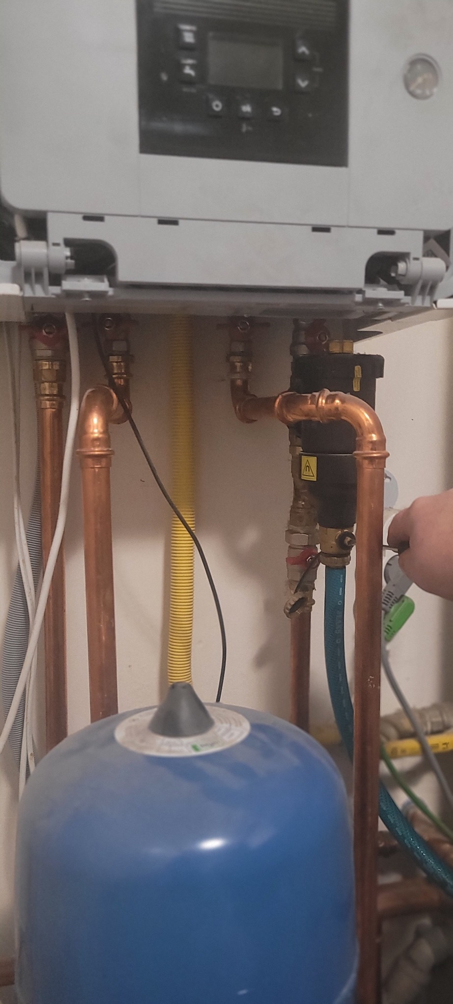 Podłączenie pieca gazowego: miedziane rury, żółty przewód gazowy, niebieski zbiornik wyrównawczy. Widoczny fragment dłoni podczas pracy.