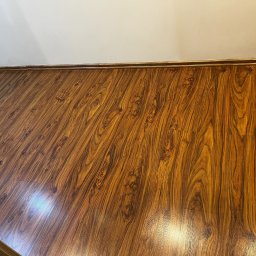 Pogotowie Stolarskie WoodCraft Marcin Krawczyk - Ukończona podłoga z paneli o wzorze drewna, z widocznymi listwami przypodłogowymi i fragmentem ściany. Układanie paneli z dbałością o detale.