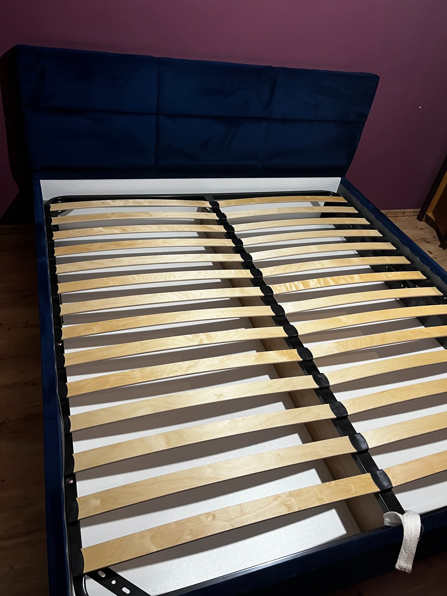Złożone łóżko tapicerowane z drewnianym stelażem i ciemnoniebieskim obiciem, widoczne z perspektywy. Elementy ramy połączone czarnymi łącznikami.