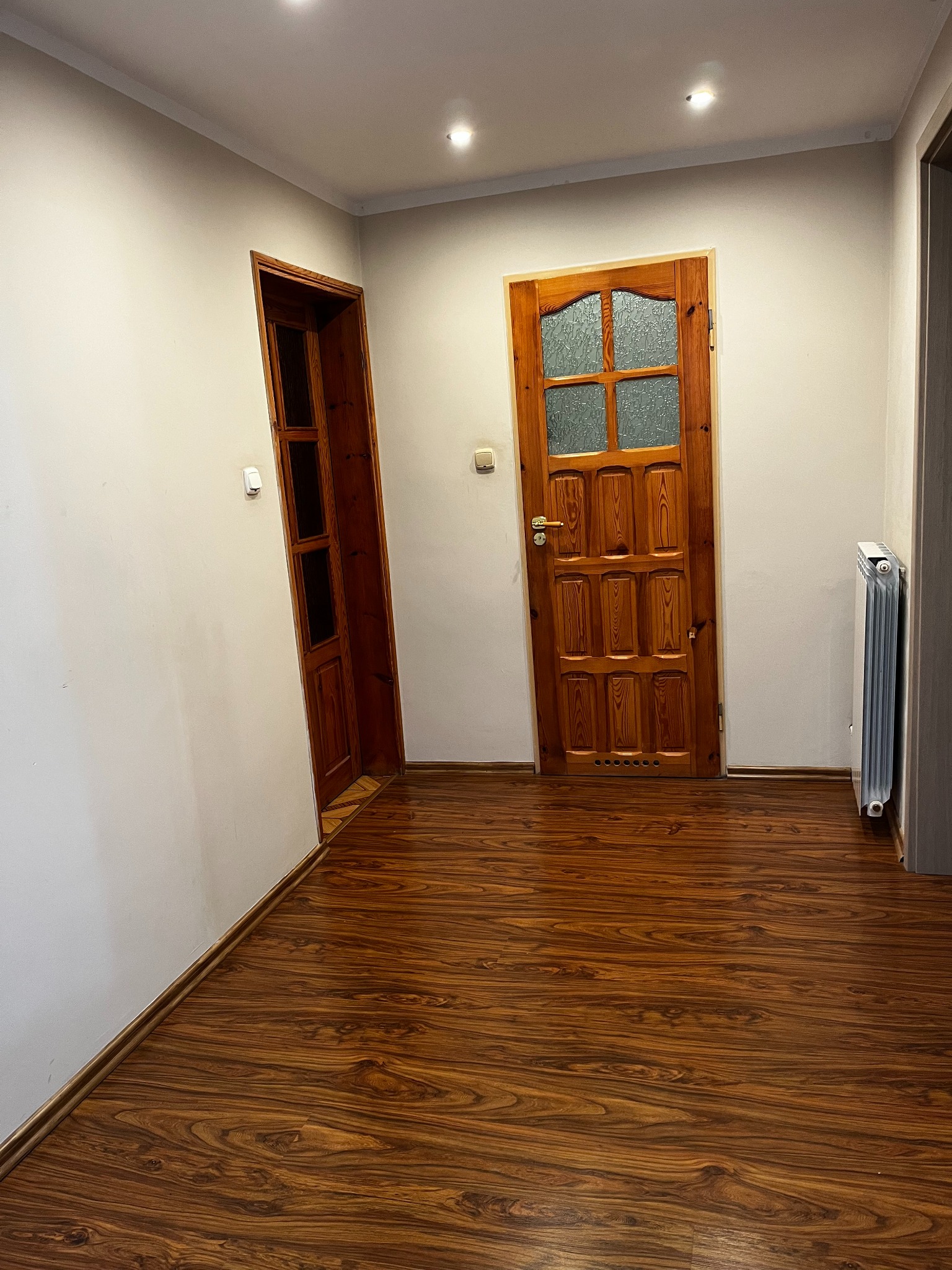 Korytarz z panelami podłogowymi o wzorze drewna, widoczne dwie drewniane ościeżnice i drzwi. Grzejnik przy ścianie. Jasne ściany i sufit z oświetleniem punktowym.