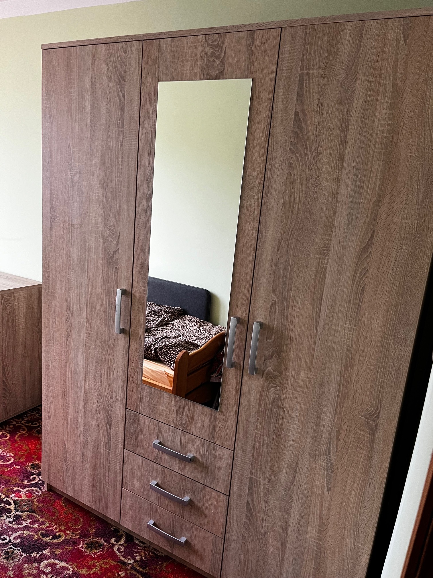 Szafa trzydrzwiowa z lustrem, wykończona w okleinie imitującej drewno. Widoczne metalowe uchwyty i trzy szuflady. W lustrze odbija się fragment sypialni.