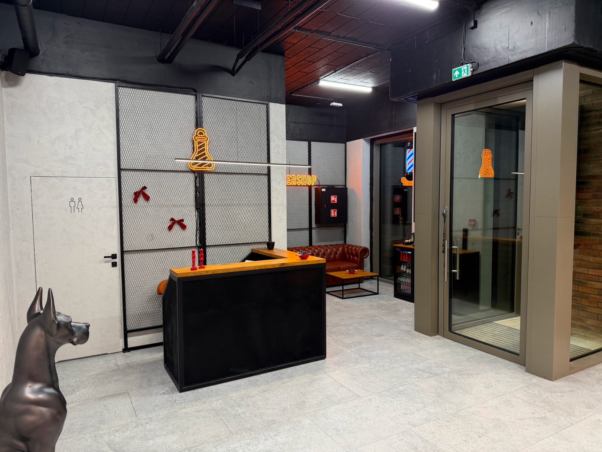 Nowoczesne wnętrze salonu z recepcją, neonem w kształcie maszynki i rzeźbą psa. Widoczna poczekalnia z brązową sofą i przeszklona winda.