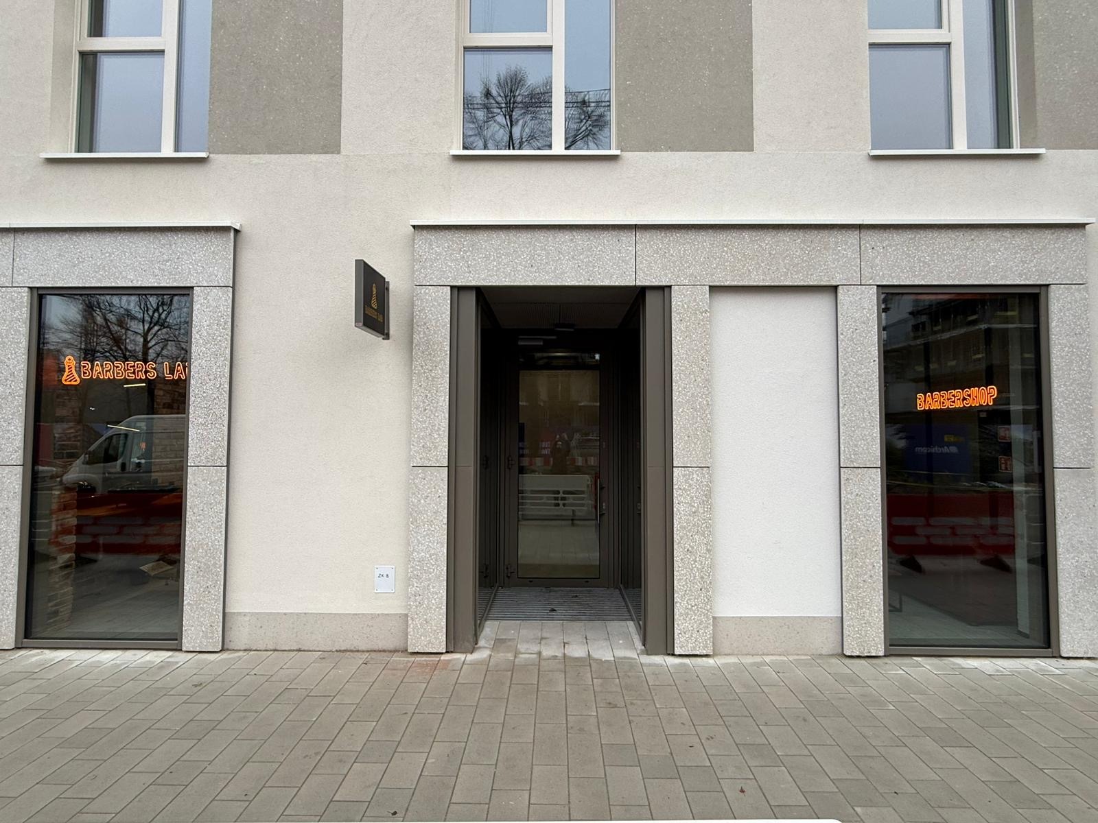 Fasada budynku z wejściem do salonu z neonami 'Barbers Lab' i 'Barbershop' po bokach. Widoczne okna i chodnik z kostki.