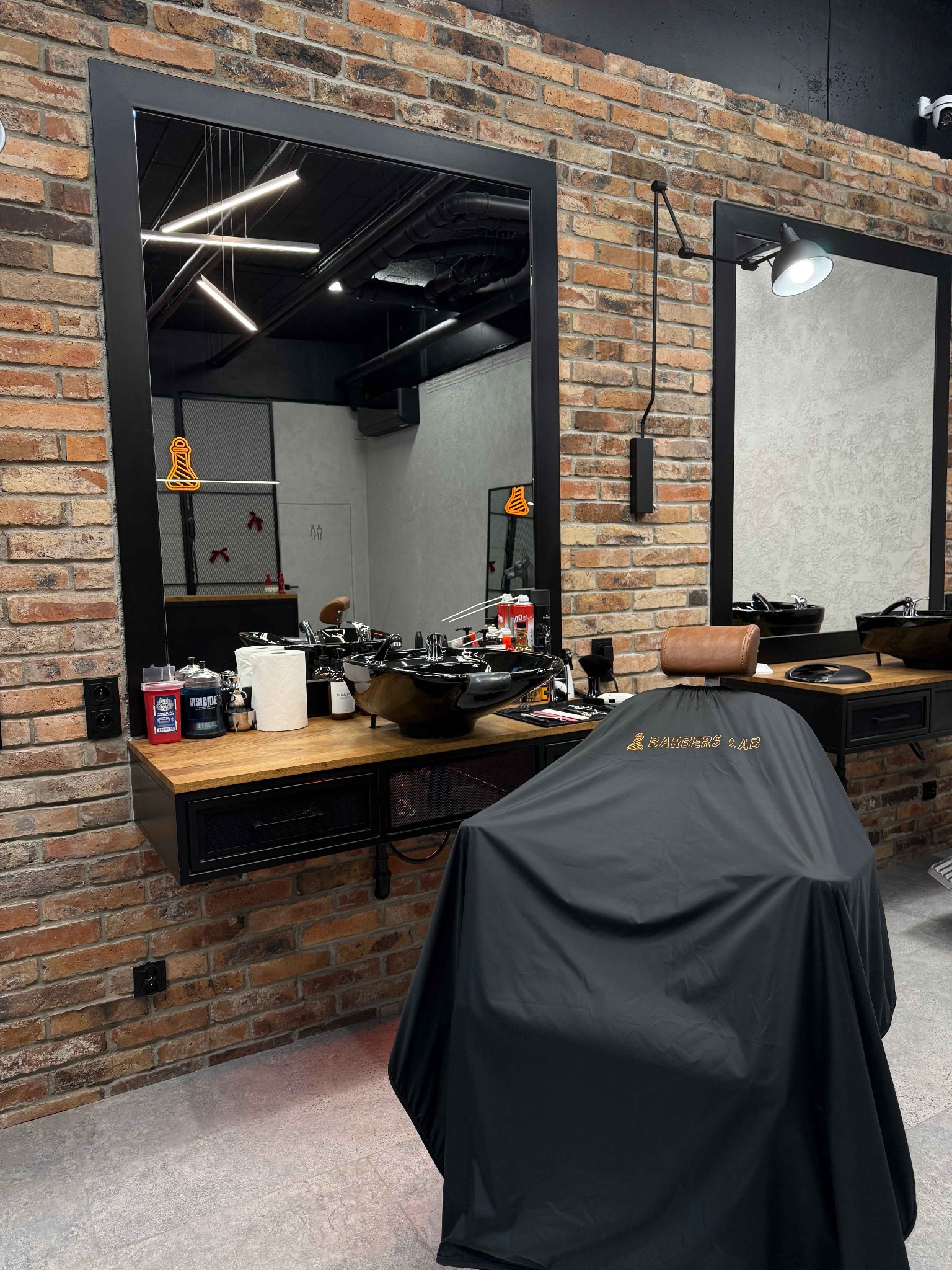 Nowoczesne wnętrze salonu z ceglaną ścianą, dużym lustrem i fotelem fryzjerskim z peleryną 'Barbers Lab'. Widoczne akcesoria i oświetlenie.