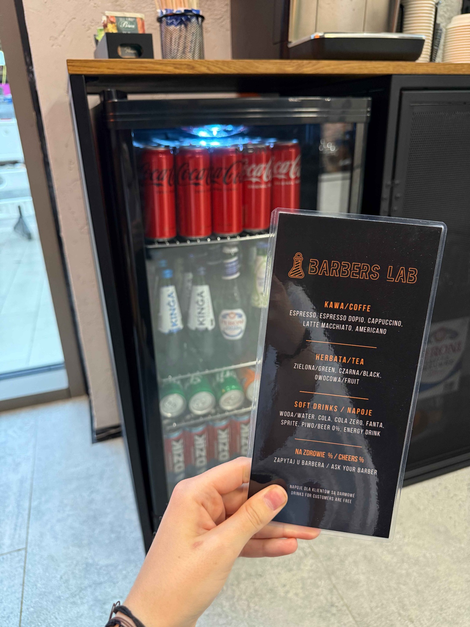 Menu napojów w salonie barberskim, trzymane w dłoni na tle lodówki z napojami. Logo 'Barbers Lab' widoczne na karcie menu.