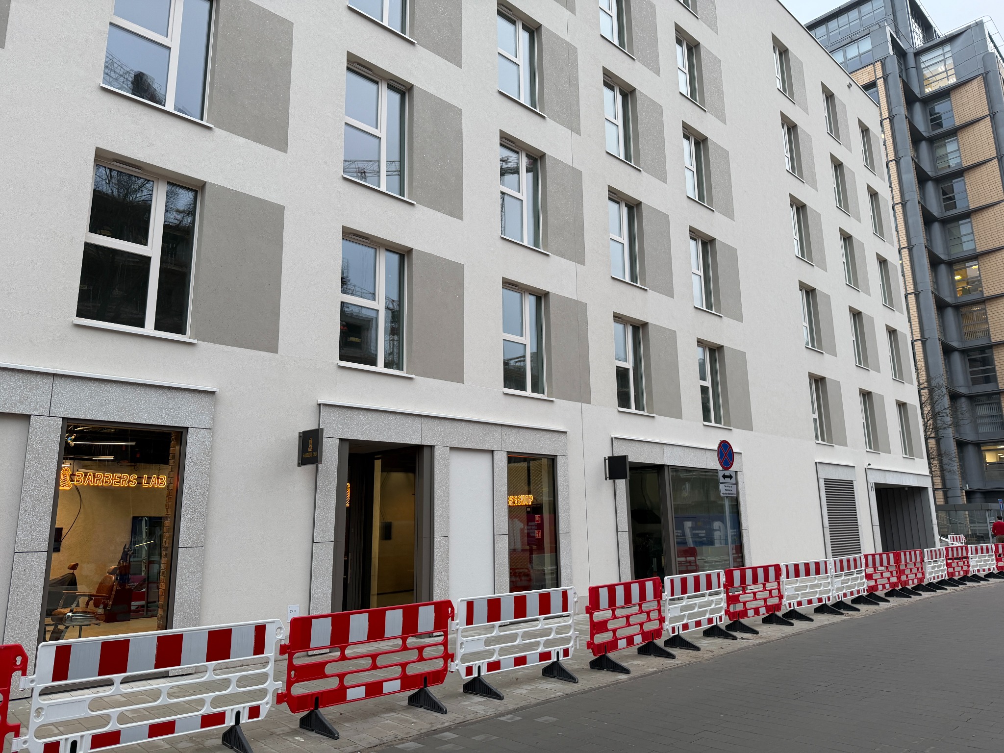 Fasada budynku z salonami fryzjerskimi 'Barbers Lab' i 'Barbershop', oddzielona od chodnika barierami ochronnymi w kolorze czerwono-białym.