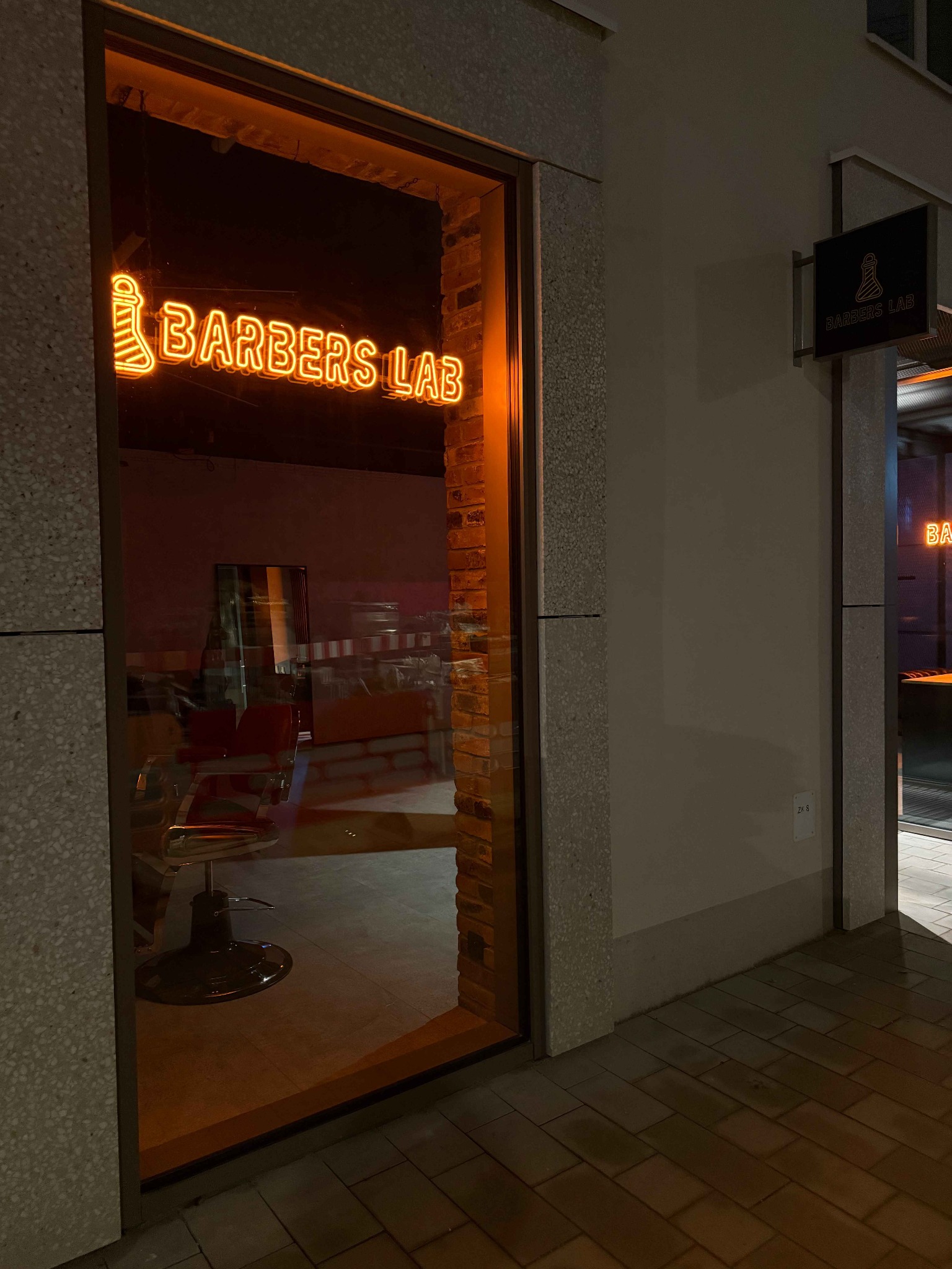 Neonowy szyld 'Barbers Lab' nad wejściem do salonu, widoczny fotel fryzjerski wewnątrz. Dodatkowy szyld na ścianie budynku. Ujęcie nocne.
