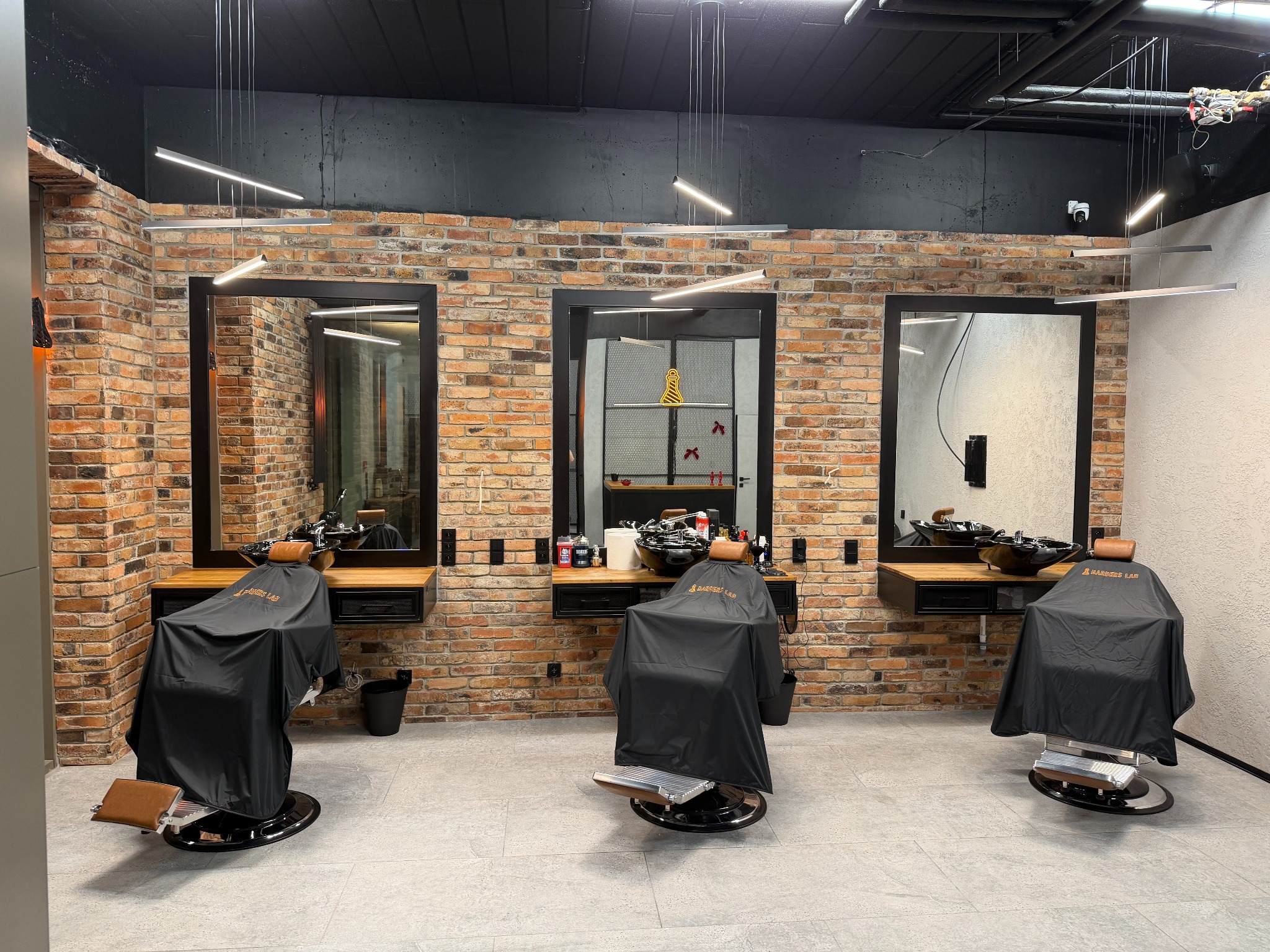 Nowoczesne stanowiska w salonie: trzy fotele z pelerynami, lustra w czarnych ramach, ceglana ściana, minimalistyczne oświetlenie. Widok na wnętrze salonu.