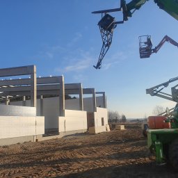 Meno - Montaż płyt warstwowych na hali w trakcie budowy. Widoczne płyty, betonowe słupy i dźwig teleskopowy Merlo Roto. Słoneczny dzień, błękitne niebo.