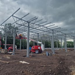 Meno - Stalowa konstrukcja hali w trakcie montażu, widoczne podnośniki koszowe i pracownicy. Pochmurne niebo w tle. Widok z poziomu gruntu.