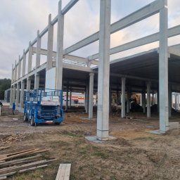 Meno - Konstrukcja hali w Siedlcach, widok z zewnątrz. Elementy betonowe, podnośnik koszowy, przygotowane płyty warstwowe. Etap budowy, surowy stan.