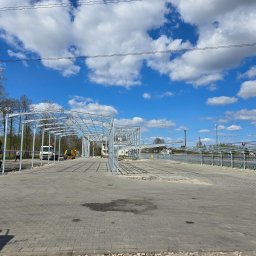 Meno - Stalowa konstrukcja hali w trakcie budowy, widoczne maszyny budowlane, błękitne niebo z chmurami w tle. Ujęcie z poziomu gruntu, szeroki kadr.