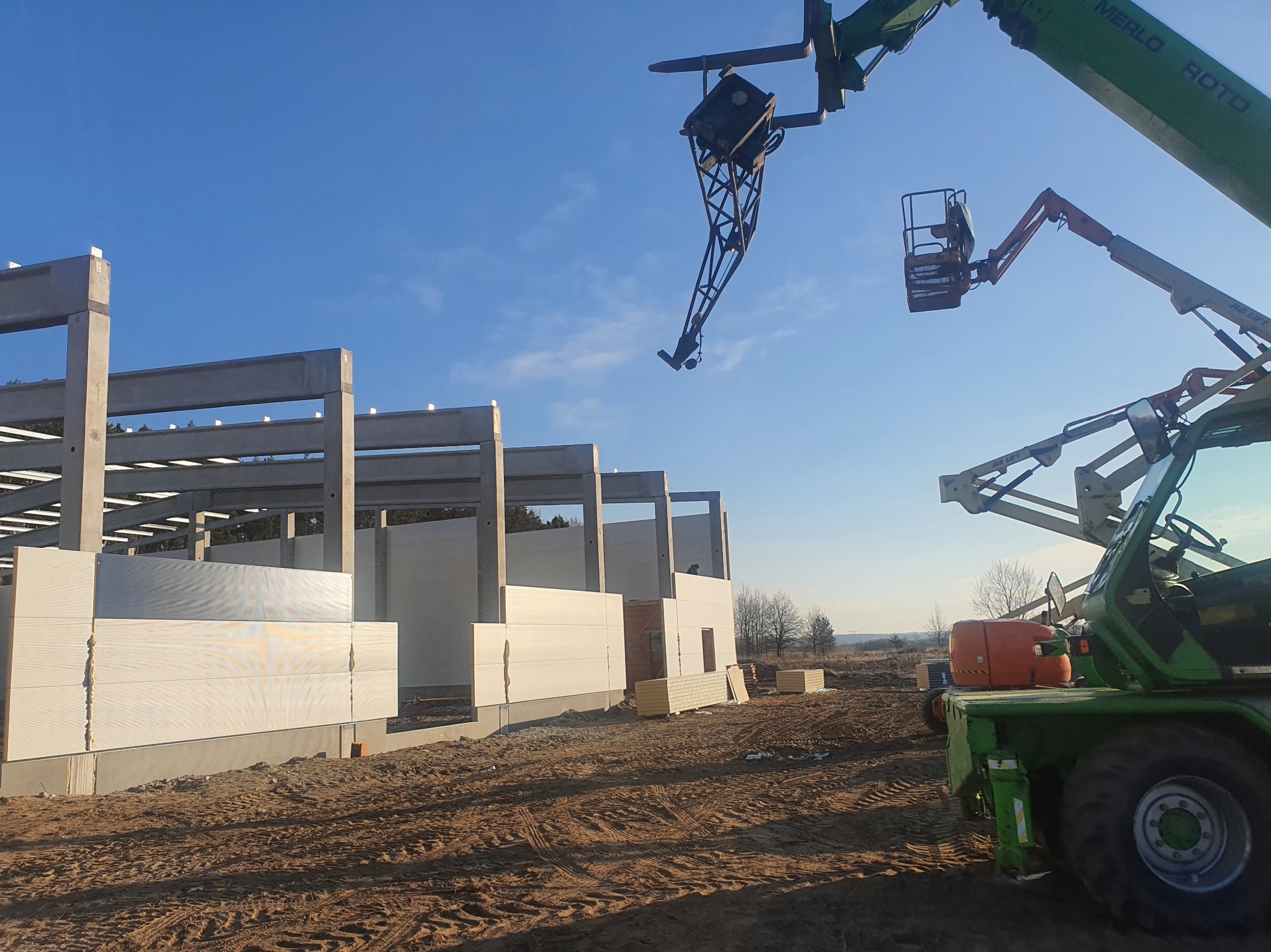 Montaż płyt warstwowych na hali w trakcie budowy. Widoczne płyty, betonowe słupy i dźwig teleskopowy Merlo Roto. Słoneczny dzień, błękitne niebo.