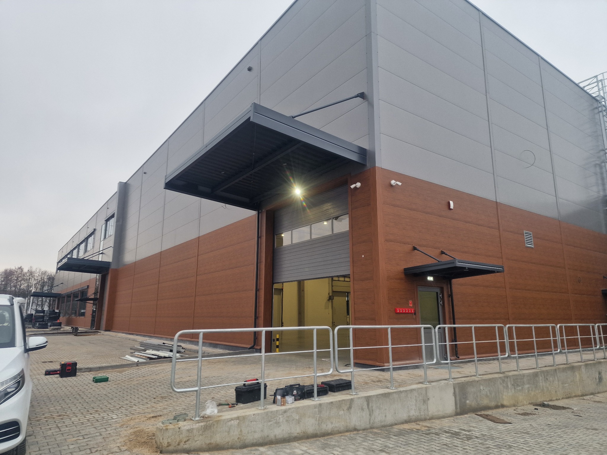 Nowoczesna hala magazynowa z szarymi panelami i brązową elewacją. Widoczne narzędzia i elementy montażowe sugerują etap końcowy budowy. Jasne oświetlenie i barierki.