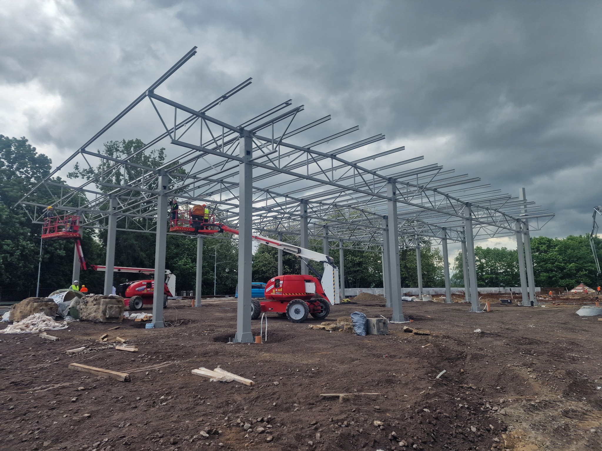 Stalowa konstrukcja hali w trakcie montażu, widoczne podnośniki koszowe i pracownicy. Pochmurne niebo w tle. Widok z poziomu gruntu.