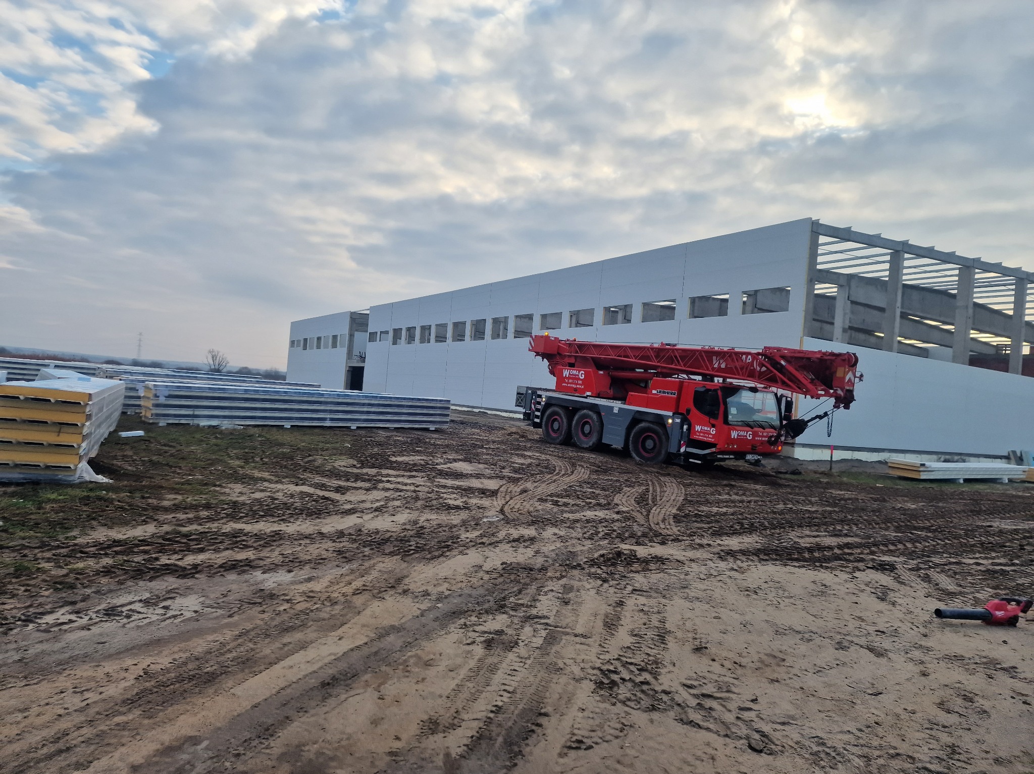 Budowa hali z widocznym dźwigiem i stertą płyt warstwowych. Częściowo ukończona konstrukcja stalowa z białymi panelami. Teren budowy z błotem i śladami opon.