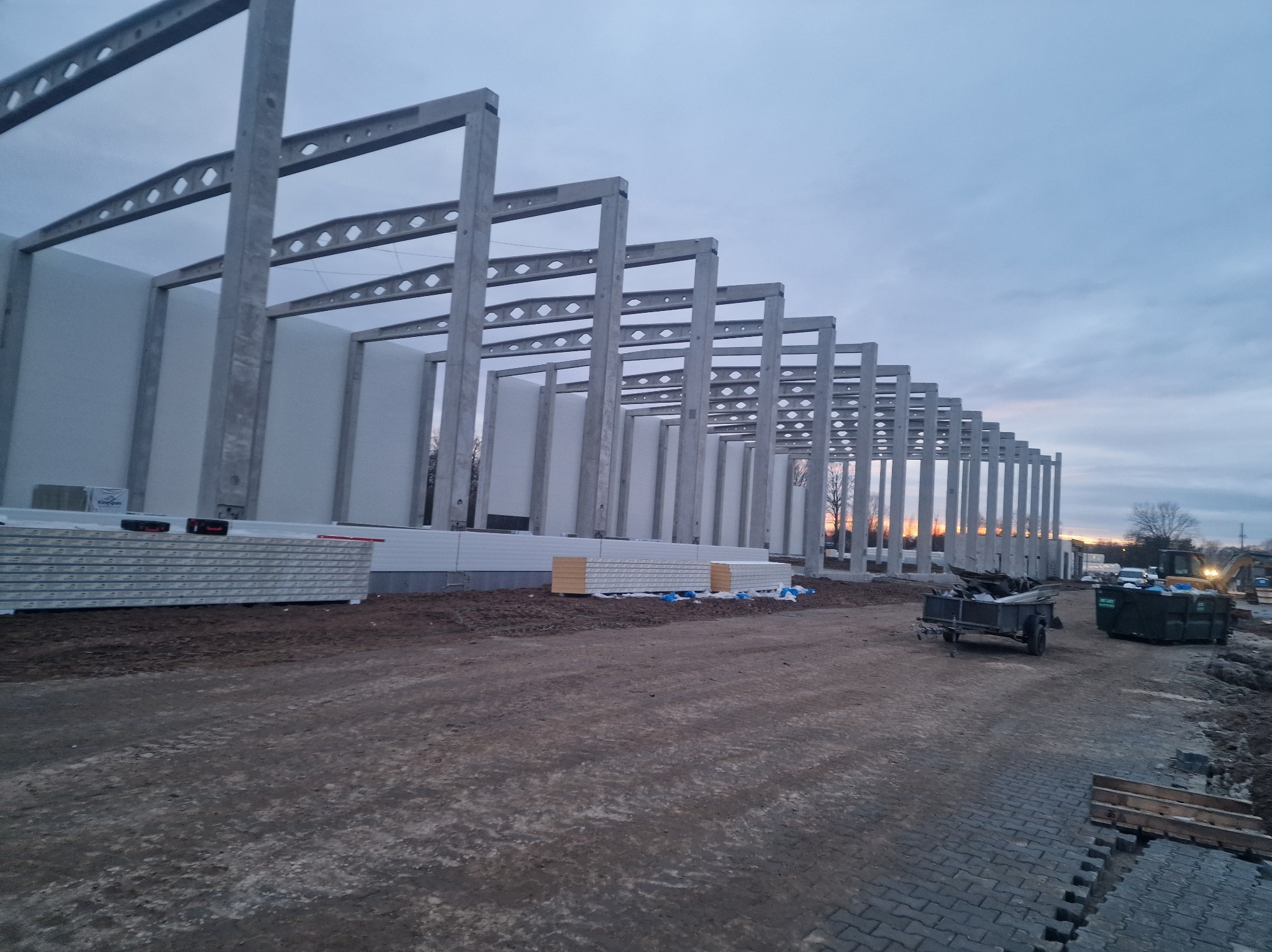 Wznoszenie hali: betonowy szkielet konstrukcyjny z widocznymi panelami warstwowymi, przygotowanymi do montażu. W tle zachodzące słońce.