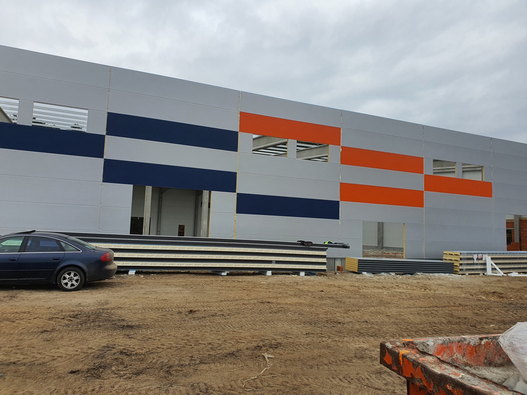 Hala w budowie z kolorowymi panelami (szary, granatowy, pomarańczowy). Na pierwszym planie stos płyt warstwowych i zaparkowany samochód osobowy.