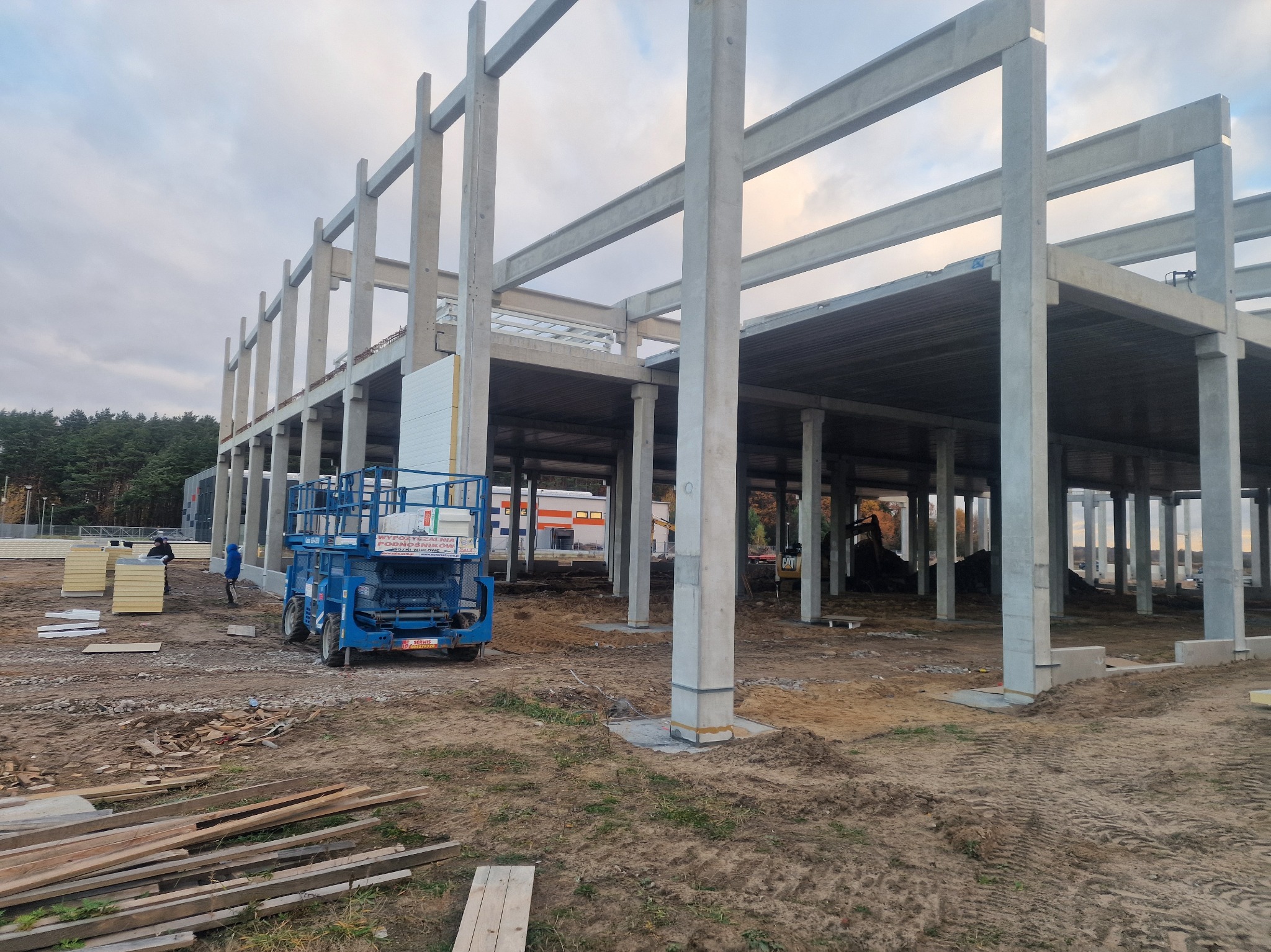 Konstrukcja hali w Siedlcach, widok z zewnątrz. Elementy betonowe, podnośnik koszowy, przygotowane płyty warstwowe. Etap budowy, surowy stan.