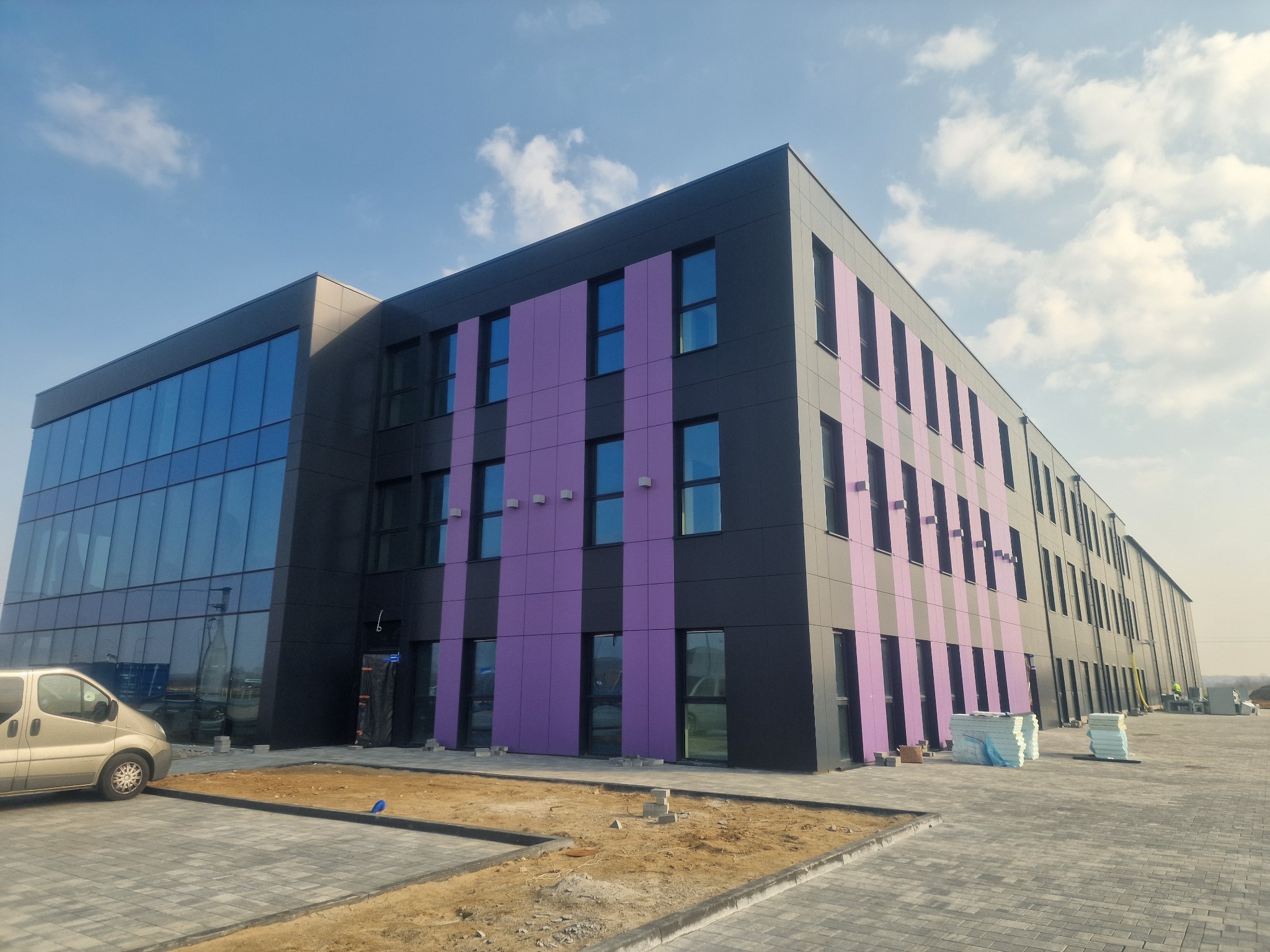 Nowoczesna hala z panelami w kolorze grafitu i fioletu, duże okna, kostka brukowa. Widoczny fragment samochodu dostawczego i materiały budowlane. Błękitne niebo w tle.