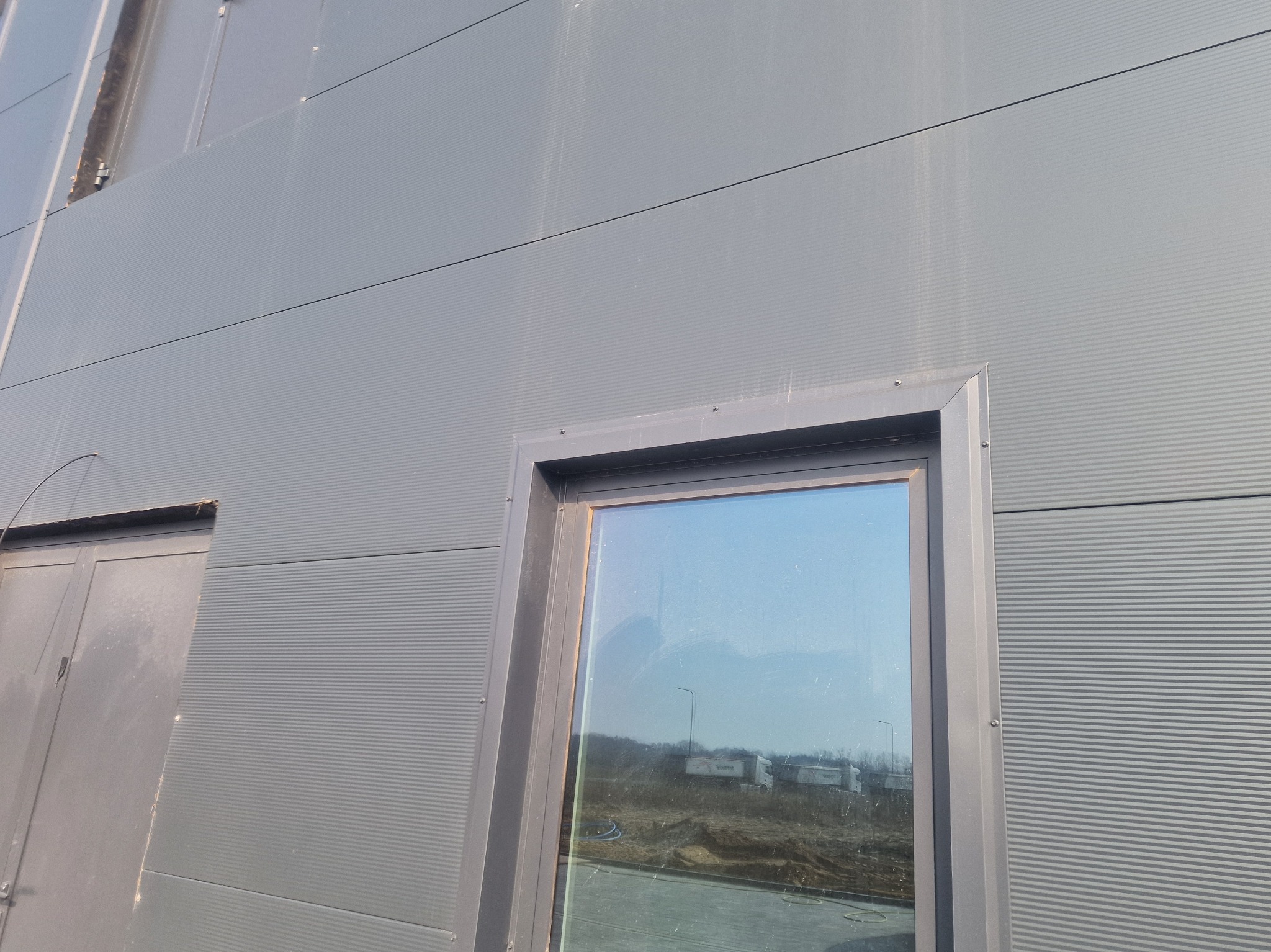 Szara ściana hali z płyty warstwowej z oknem i drzwiami. Widoczne łączenia płyt. W oknie odbija się krajobraz z ciężarówkami. Ujęcie z dołu.