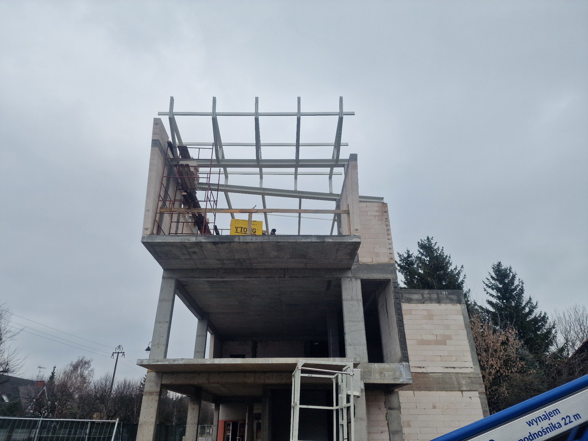 Wznoszenie dwukondygnacyjnej konstrukcji z betonu komórkowego Ytong, z widocznym szkieletem dachu i elementami rusztowania. Ujęcie z dołu na tle pochmurnego nieba.