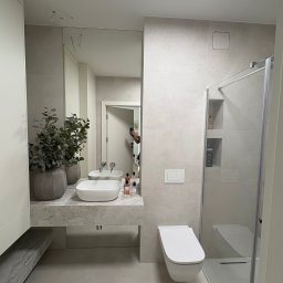 Rasz - Jasna łazienka z kabiną prysznicową, umywalką na blacie z rośliną i podwieszaną toaletą. Minimalistyczny design z szarymi płytkami i lustrem.