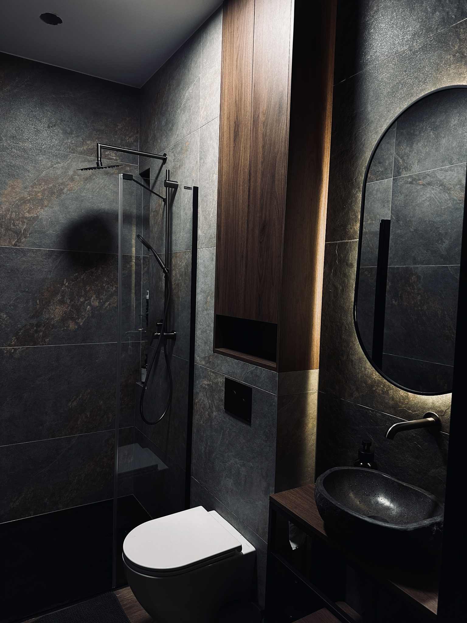Nowoczesna łazienka z ciemnymi płytkami imitującymi kamień, kabiną prysznicową typu walk-in i owalnym lustrem z podświetleniem LED. Umywalka kamienna.