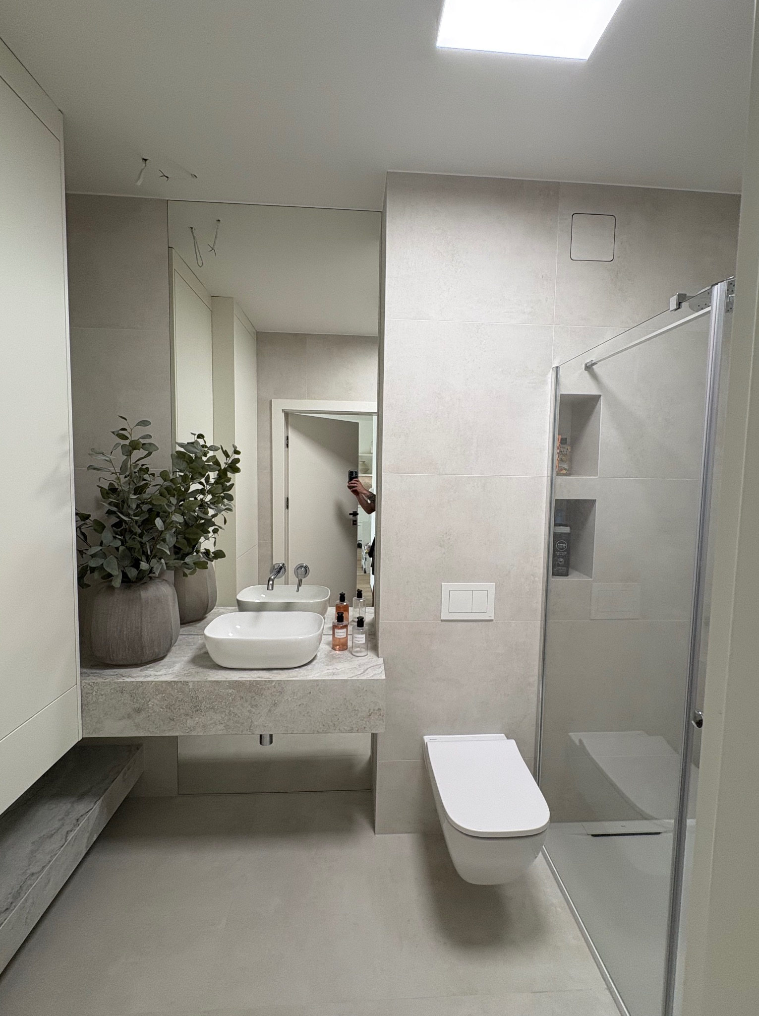 Jasna łazienka z kabiną prysznicową, umywalką na blacie z rośliną i podwieszaną toaletą. Minimalistyczny design z szarymi płytkami i lustrem.